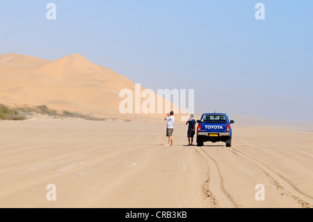 I turisti in piedi accanto a un veicolo fuoristrada a scattare foto in Sandwich Harbour, parco nazionale, parte di scheletro della Namibia Foto Stock