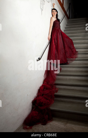 Giovane donna che indossa un abito rosso con un treno lungo stando in piedi sulla scala di pietra in un vano scala Foto Stock