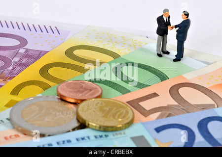 Statuine in miniatura dei dirigenti stringono le mani mentre sta in piedi sulle banconote e monete in euro, immagine simbolica per il business Foto Stock
