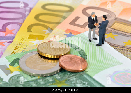 Statuine in miniatura dei dirigenti stringono le mani mentre sta in piedi sulle banconote e monete in euro, immagine simbolica per il business Foto Stock