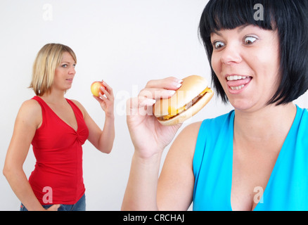 Due donne mangiare hamburger e Apple, con la donna bionda con Apple in background, cercando enviously alla donna con il burger Foto Stock