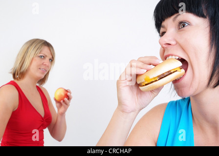 Due donne mangiare hamburger e Apple, con la donna bionda con Apple in background, cercando enviously alla donna con il burger Foto Stock