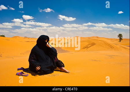 Un anziano Berber donna che indossa abiti neri seduta a piedi nudi nelle dune di sabbia, Erg Chebbi, Sahara, sud del Marocco, Marocco Foto Stock