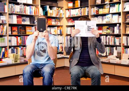 Uomo in possesso di un libro elettronico o e-book reader davanti al suo volto seduto accanto a un altro uomo in possesso di un libro convenzionale in Foto Stock