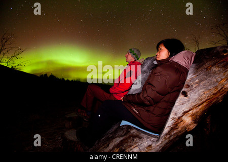 Coppia giovane, donna e uomo seduto in una sedia in legno, visualizzazione, guardando il verde polare settentrionale luci, Aurora Boreale Foto Stock