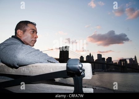 Riflessivo African American businessman rilassante su banco, ponte di Brooklyn e gli edifici della città in background Foto Stock