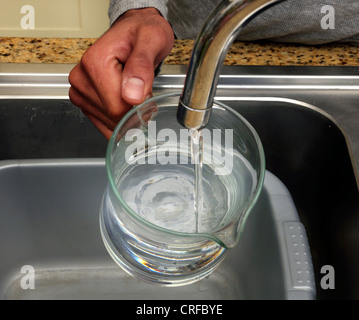 Afro-caraibica mani dell'uomo il riempimento di acqua in un bicchiere di vetro da un rubinetto Foto Stock