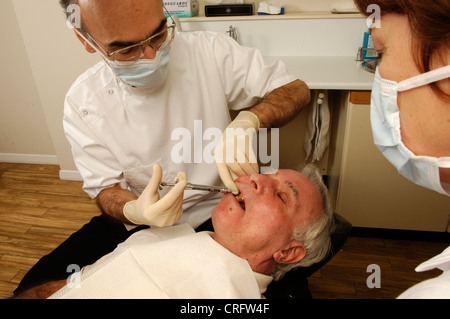Guardati dal suo assistente, un dentista inietta un dolore locale uccidendo la sostanza anestetica nel paziente un gum per intorpidire la zona circostante prima di iniziare una procedura dentale. Foto Stock