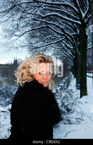 Donna bionda nel mantello nero in piedi nella neve Foto Stock