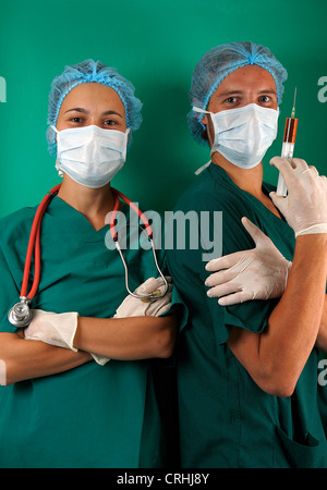 Due chirurghi uno con uno stetoscopio uno con una siringa riempita con liquido rosso sia con maschere e retine per capelli. Sfondo verde. Foto Stock