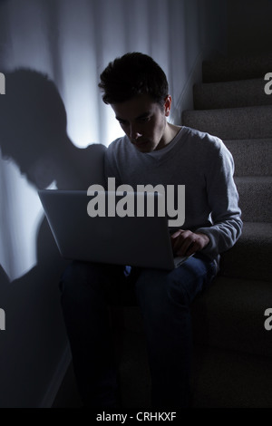 Maschio adolescente usando il suo computer portatile, seduta in scala scura.. Foto Stock