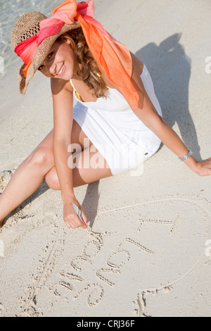 Giovane e bella donna che indossa un abito bianco e un cappello per il sole, seduto sulla spiaggia, il disegno di un cuore nella sabbia Foto Stock