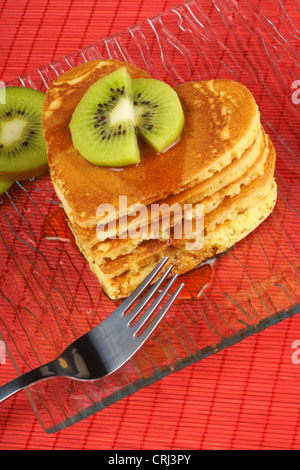 Pila di heart-shaped pancake con sciroppo e kiwi su un vetro trasparente piatto su uno sfondo rosso. Per il giorno di San Valentino Foto Stock