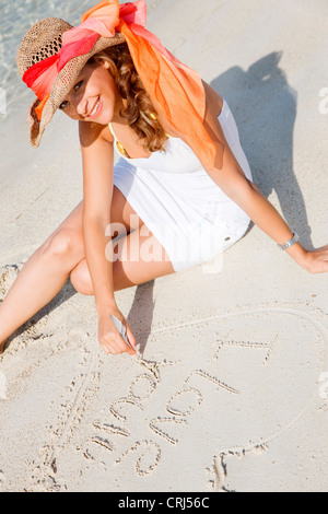 Giovane e bella donna che indossa un abito bianco e un cappello per il sole, seduto sulla spiaggia, il disegno di un cuore nella sabbia Foto Stock