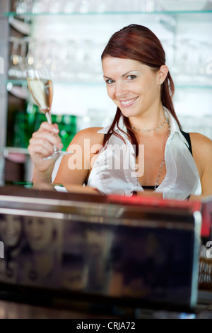 Giovane barista dietro il bar sollevando un bicchiere di vino frizzante con un sorriso Foto Stock