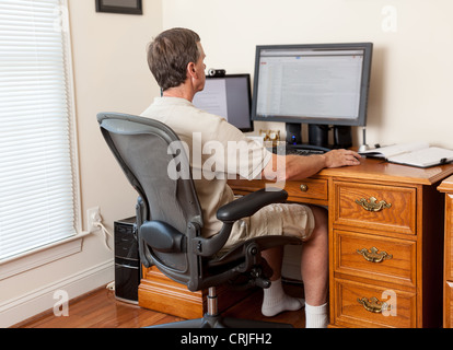 Uomo che lavora da casa ufficio in pantaloncini corti con scrivania con due monitor Foto Stock