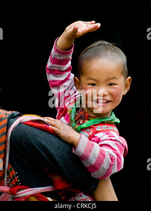 Un felice giovani tibetani toddler cinese le onde a la telecamera mentre sulla sua schiena delle madri. Foto Stock