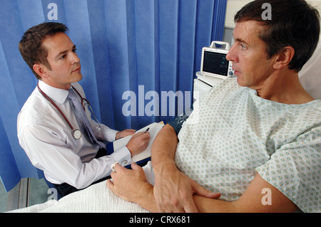 Un medico sulla sua ward round si siede e parla con un paziente di sesso maschile al suo posto letto ospedaliero. Foto Stock