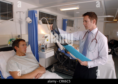 Un medico sulla sua ward round recensioni un x-ray durante un ospedale di consultazione al posto letto con la sua paziente di sesso maschile. Foto Stock