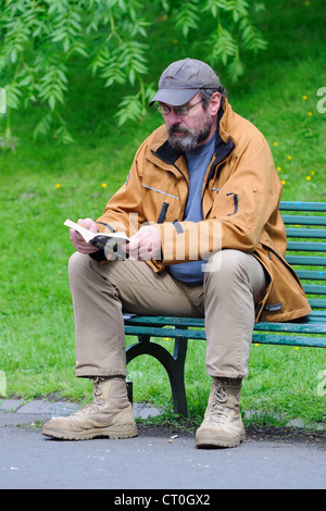 Un uomo trascorre il tempo in tranquillità seduti su una panchina nel parco la lettura di un libro tascabile. Foto Stock
