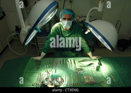 Un chirurgo mascherato appoggiato su di una tavola di sterili strumenti chirurgici in un teatro ospedaliero. Foto Stock