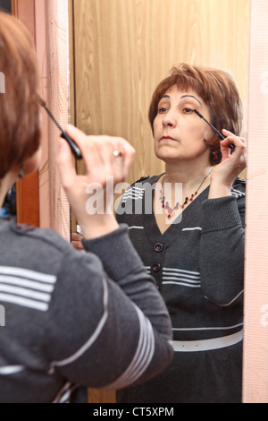 Senior donna caucasica applicare il make-up contro uno specchio in ambiente domestico Foto Stock