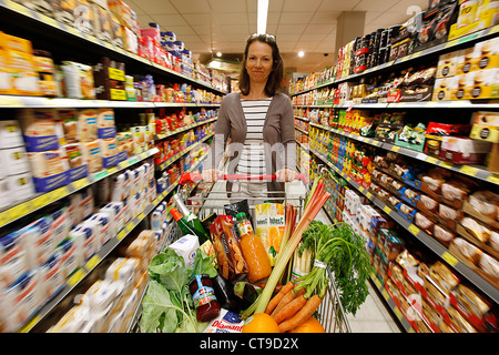 Grande supermercato, donna spinge un intero carrello di shopping lungo i piani di appoggio con prodotti alimentari. Foto Stock