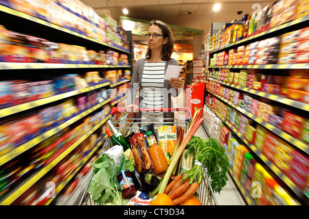 Grande supermercato, donna spinge un intero carrello di shopping lungo i piani di appoggio con prodotti alimentari. Foto Stock