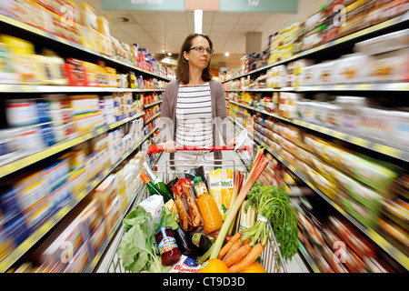 Grande supermercato, donna spinge un intero carrello di shopping lungo i piani di appoggio con prodotti alimentari. Foto Stock