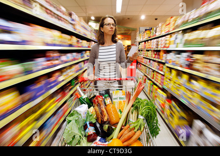 Grande supermercato, donna spinge un intero carrello di shopping lungo i piani di appoggio con prodotti alimentari. Foto Stock