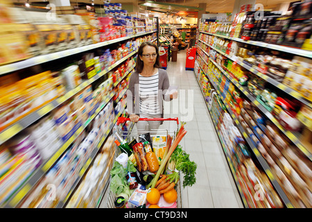 Grande supermercato, donna spinge un intero carrello di shopping lungo i piani di appoggio con prodotti alimentari. Foto Stock