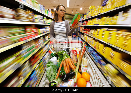 Grande supermercato, donna spinge un intero carrello di shopping lungo i piani di appoggio con prodotti alimentari. Foto Stock