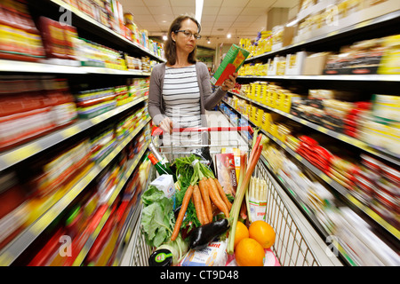 Grande supermercato, donna spinge un intero carrello di shopping lungo i piani di appoggio con prodotti alimentari. Foto Stock