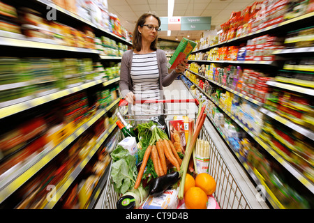 Grande supermercato, donna spinge un intero carrello di shopping lungo i piani di appoggio con prodotti alimentari. Foto Stock