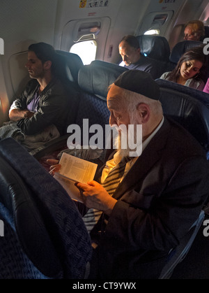 Indossando un yarmulke zucchetto, un barbuto ebreo ortodosso legge da un libro in ebraico su un aereo di linea di passeggeri durante il sonno. Foto Stock