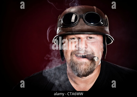 Biker in un casco fuma un sigaro su sfondo scuro Foto Stock