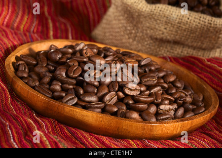 Caffè torrefatto in grani sul piatto di legno sul panno rosso con sacchetto in iuta nella parte posteriore Foto Stock