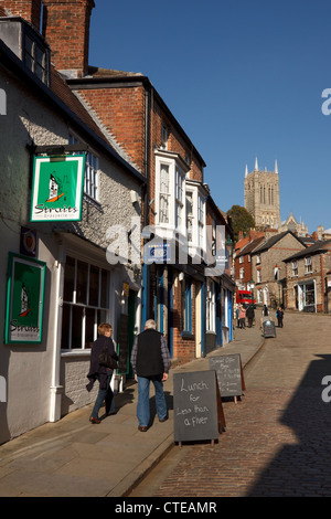 Il vecchio tradizionale negozio terrazzati fronti sulla strada di ciottoli con il duomo sopra, ripida collina, Lincoln, Lincolnshire, England, Regno Unito Foto Stock