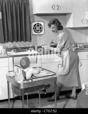 Anni Cinquanta donna madre nella cucina di casa pentola di cottura su stufa a sorridere al bambino il bambino nel seggiolino Foto Stock