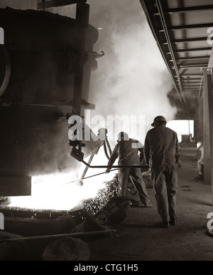 Anni sessanta COPPIA DI LAVORATORI SIDERURGICI INDOSSARE COPRICAPI RIGIDI AGITAZIONE & addensarsi di metallo fuso da colare dal crogiolo Foto Stock