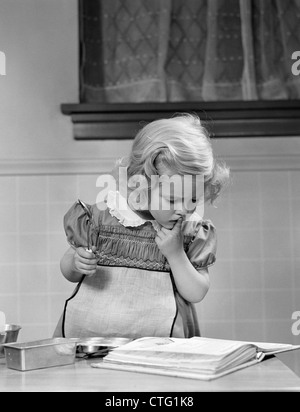 1940s bambina giocando in cucina che indossa il grembiule cercando nel Cookbook ottenere pronto per cuocere al forno Foto Stock