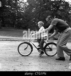 Anni sessanta padre dando figlio sulla moto un insegnamento di spinta a lui come a guidare la bicicletta Foto Stock