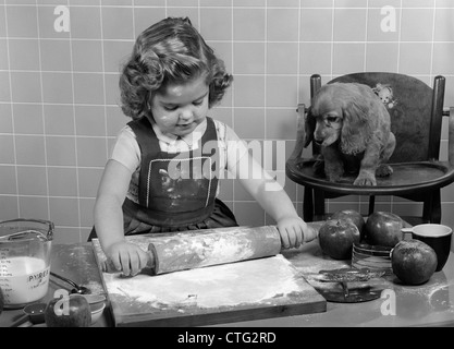 Anni Cinquanta BAMBINA ROLLING OUT APPLE base di pastafrolla per torta sul tavolo da cucina con cocker spaniel cucciolo guardando Foto Stock