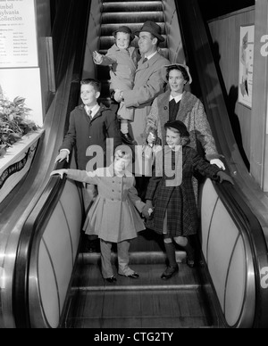 Anni Cinquanta sorridente FAMIGLIA DI 6 IN cappotti invernali SCENDENDO STORE ESCALATOR Foto Stock