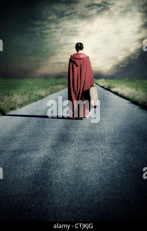 Una donna in un mantello rosso è a piedi lungo una strada con una valigia Foto Stock