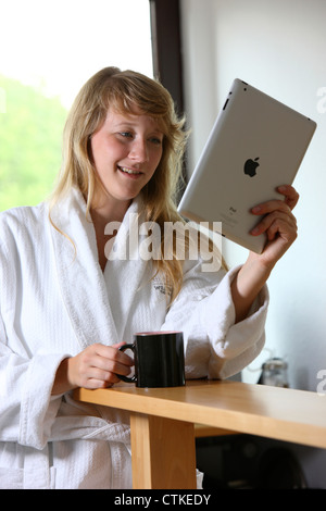 Giovane donna è utilizzando un IPad, tablet PC, la navigazione in Internet tramite connessione wireless, al mattino, avente una tazza di caffè Foto Stock