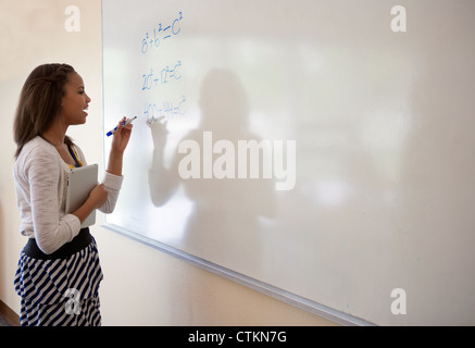 Tredici anni di African American Girl scrivere un problema di matematica a bordo in aula mentre tiene un tablet ipad. Foto Stock