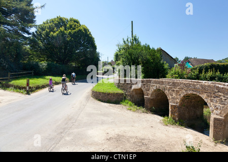 Xvii secolo packhorse Bridge crossing Dowlish Brook, con la famiglia in bicicletta attraverso il villaggio, Dowlish Wake, Somerset Foto Stock