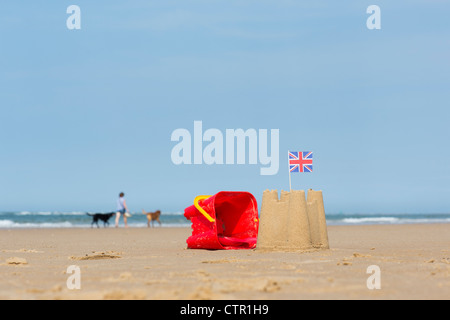 Union Jack flag in un castello di sabbia accanto a una benna childs e vanga su una spiaggia. Inghilterra Foto Stock