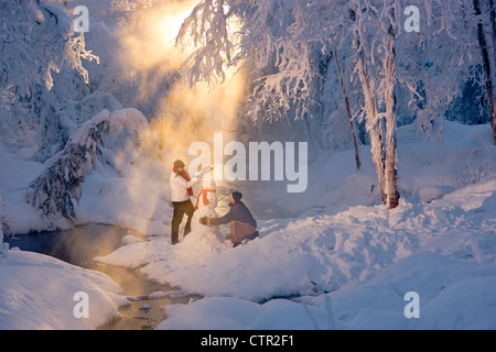 Marito e moglie la costruzione di pupazzo di neve in frosty forest retroilluminati da raggi solari del Jack russo Springs Park centromeridionale Alaska inverno Foto Stock